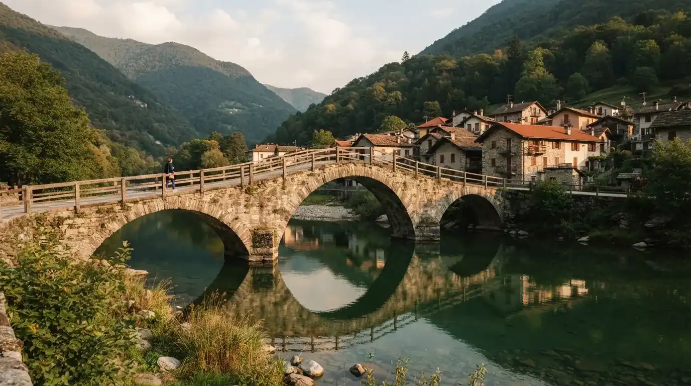 Eine alte Steinbrücke über einen ruhigen Fluss, dahinter Häuser mit Ziegeldächern und bewaldete Hügel.