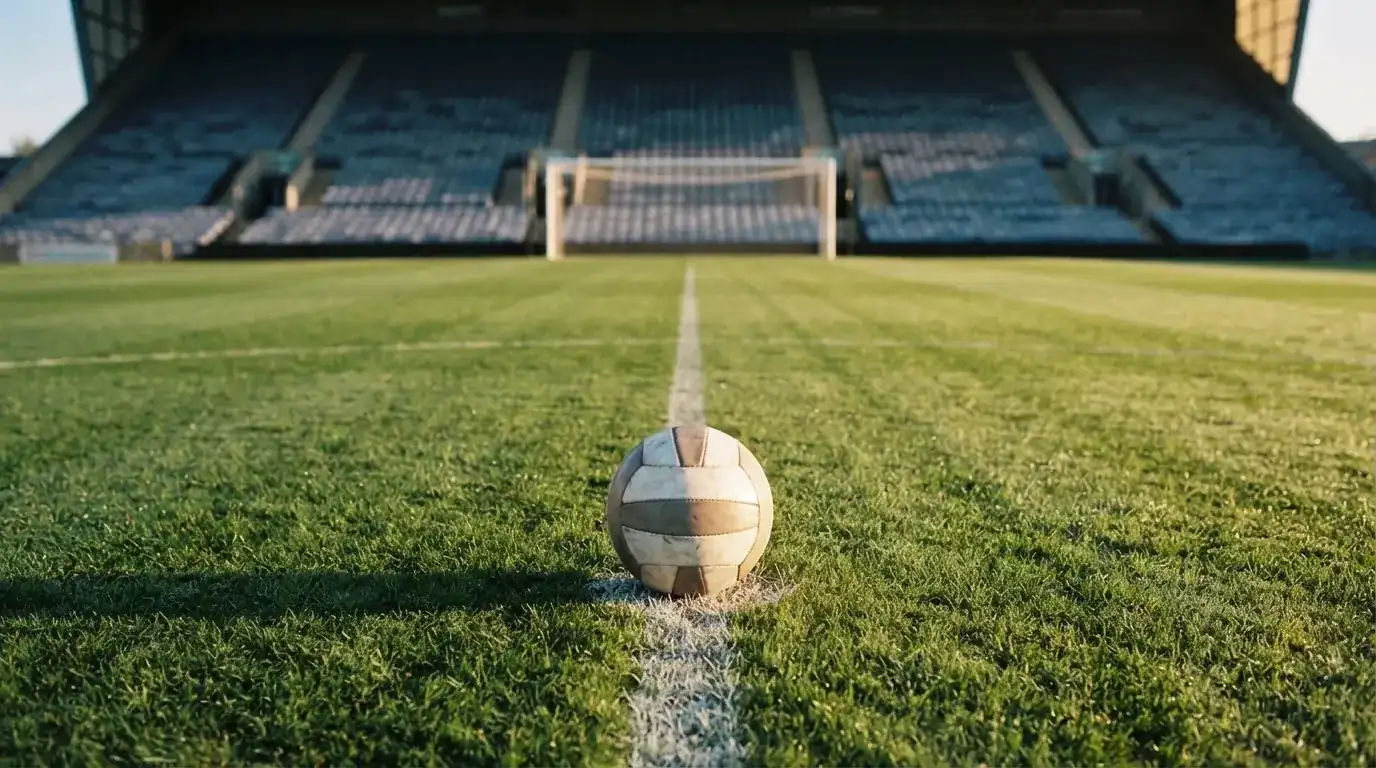 Ein Fussball liegt auf dem Anstosspunkt eines leeren Rasenplatzes im frühen Morgenlicht, dahinter erhebt sich eine menschenleere Tribüne.