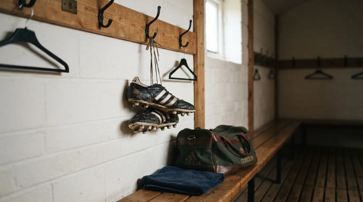 Ein Paar Fussballschuhe hängt an einem Haken in einer ruhigen Umkleide, darunter steht eine Sporttasche auf einer Holzbank.