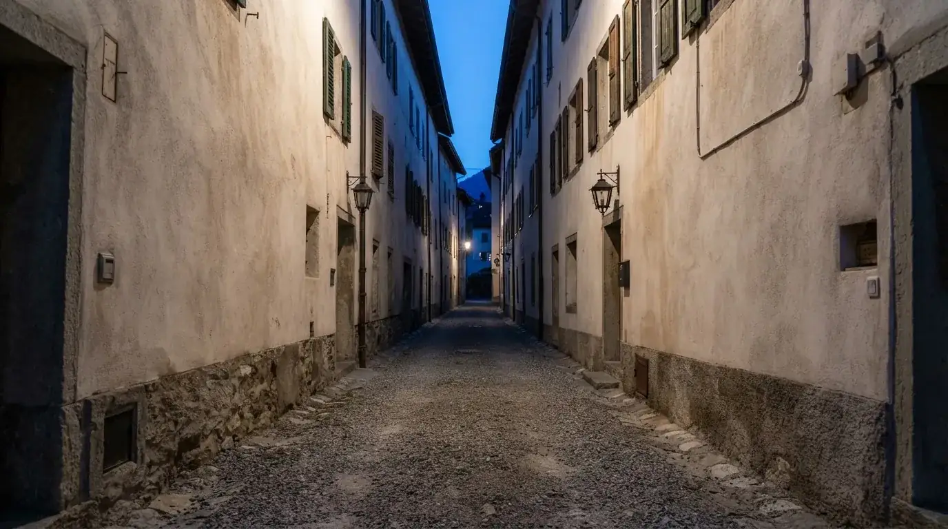 Ein schmaler Kiesweg führt zwischen zwei hellen Mauern durch eine Schweizer Altstadt, die Laternen sind noch nicht angezündet.