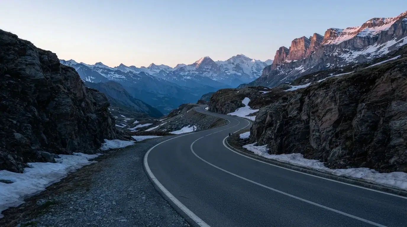 Ein Bergpass in den Schweizer Alpen mit leerer Strasse und Schneeresten am Rand im ersten Frühlicht.