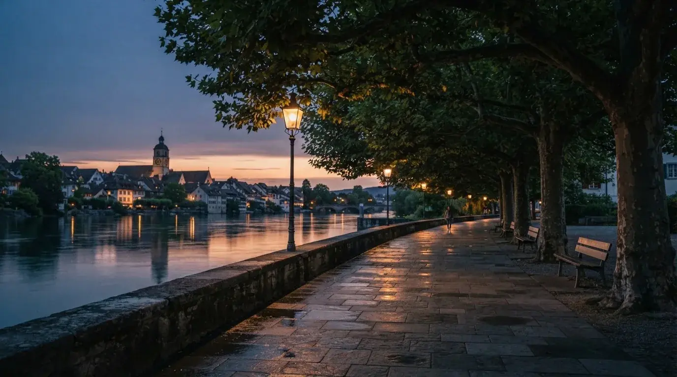 Eine Rheinpromenade mit hohen Bäumen am Abend, das Wasser spiegelt das Licht der ersten Laternen.