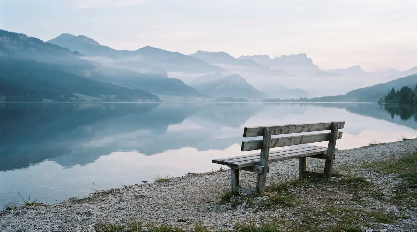 Eine Holzbank an einem Seeufer mit Blick auf eine Bergkette im Morgendunst, das Wasser ist spiegelglatt.