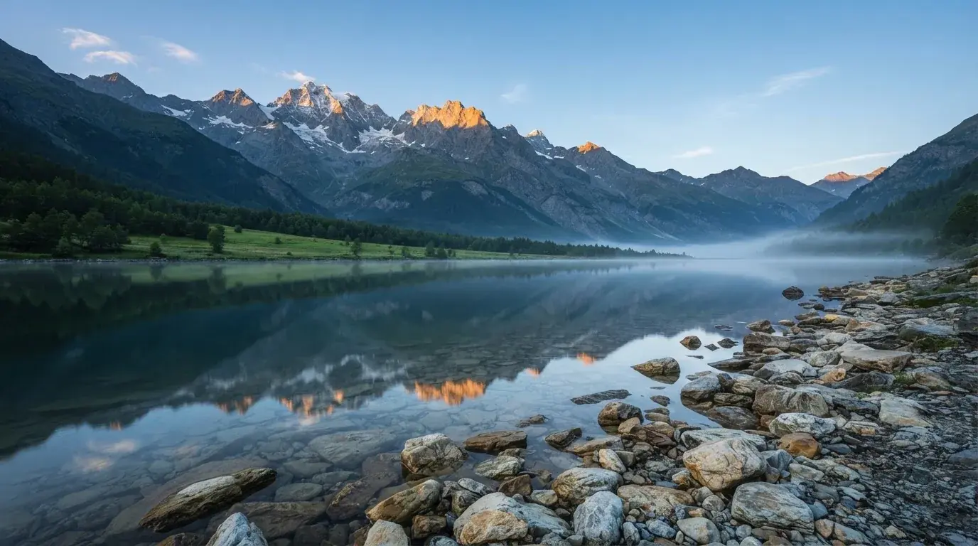 Ein Bergsee mit ruhigem Wasser und Gipfeln im Morgenlicht, am Ufer liegen glatte Steine.