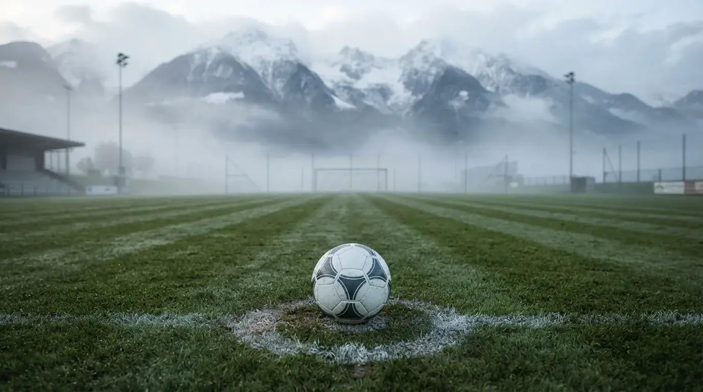 Ein einzelner Fussball auf dem Mittelpunkt eines leeren Rasens im Morgennebel, hinter dem Tor ragen Berge auf