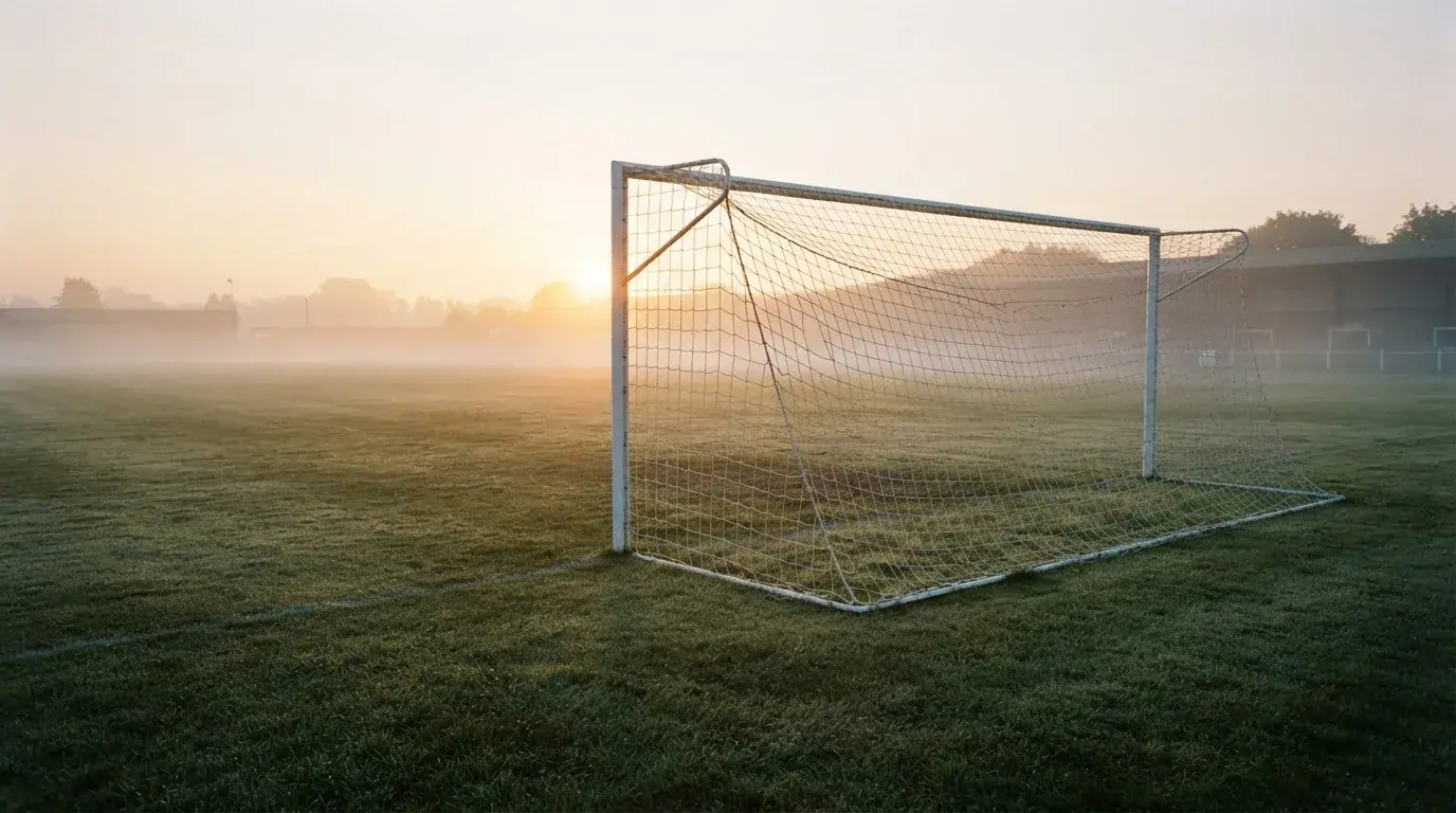 Ein Torpfosten mit Netz steht im Gegenlicht auf einem leeren Fussballplatz, über dem Rasen liegt Nebel.